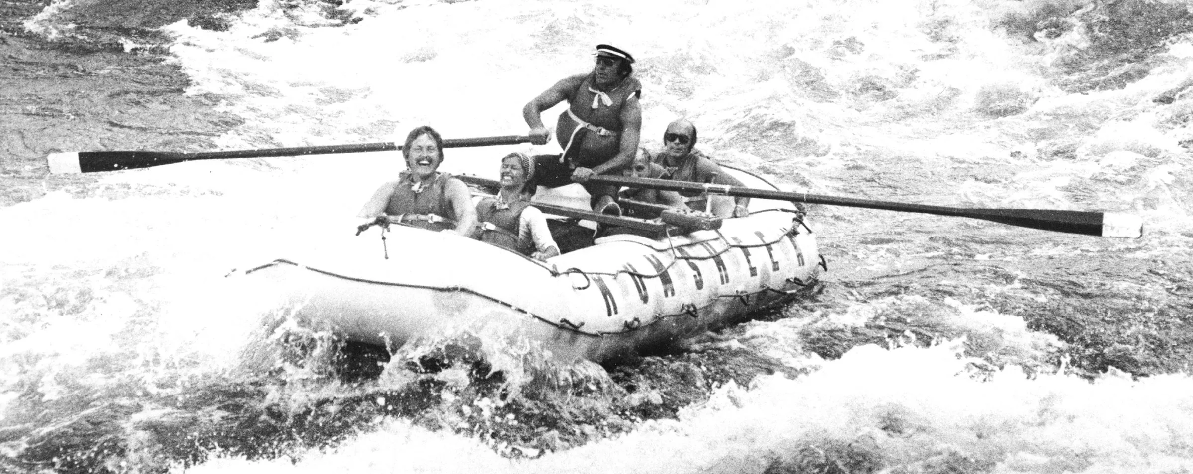 A black and white photo of a guide rowing a raft marked Kumsheen through whitewater, with three laughing passengers.