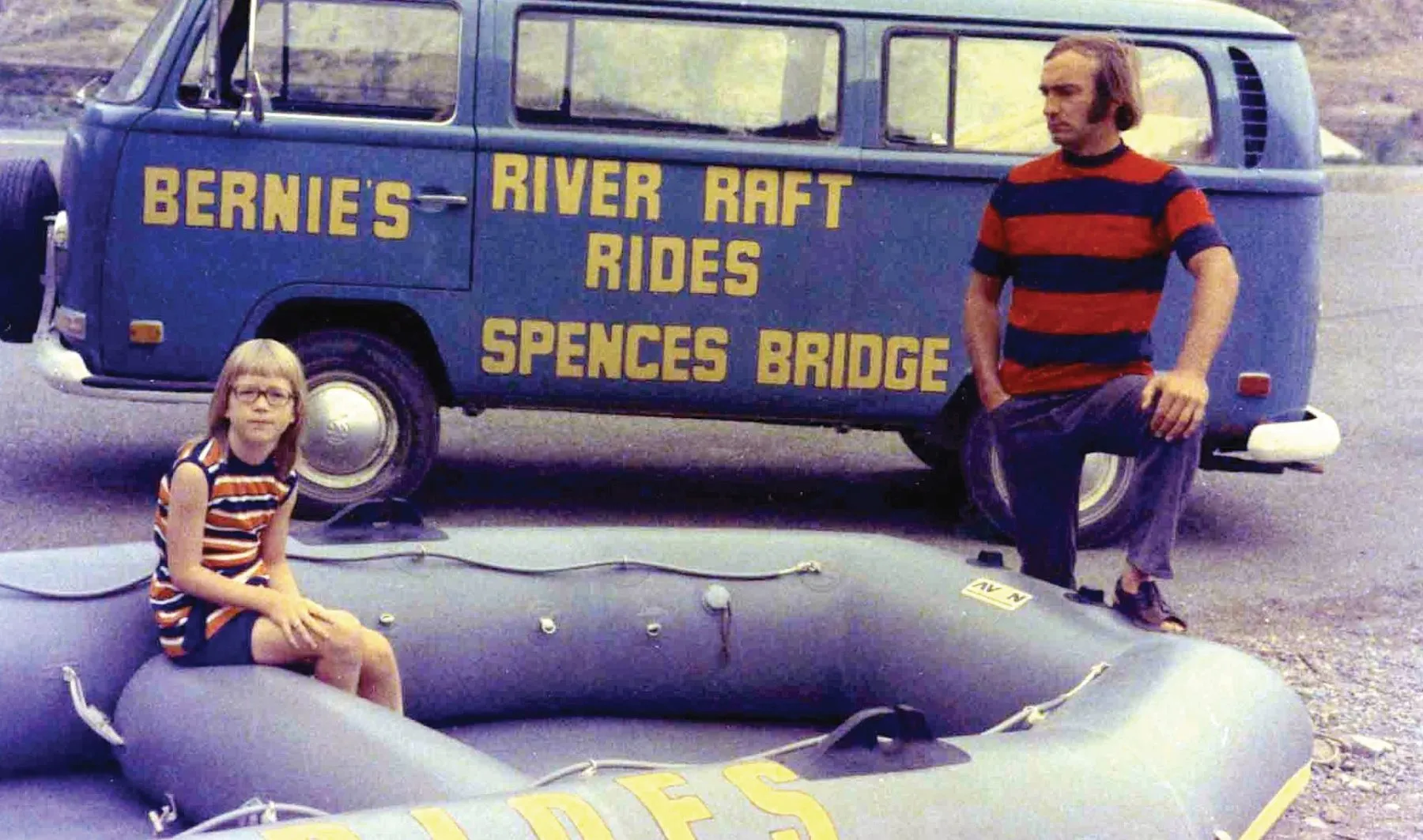 A blue Volkswagen van hand-lettered Bernie’s River Raft Rides, Spences Bridge, with a man kneeling and a child sitting on an inflatable raft in front of it.