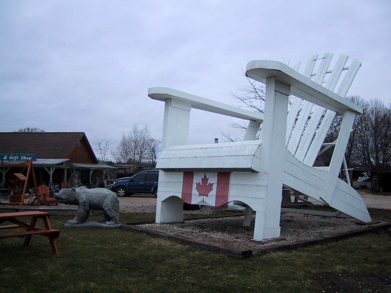 A white Muskoka chair several times the height of a person, with a Canadian flag on its front, beside a roadside shop.