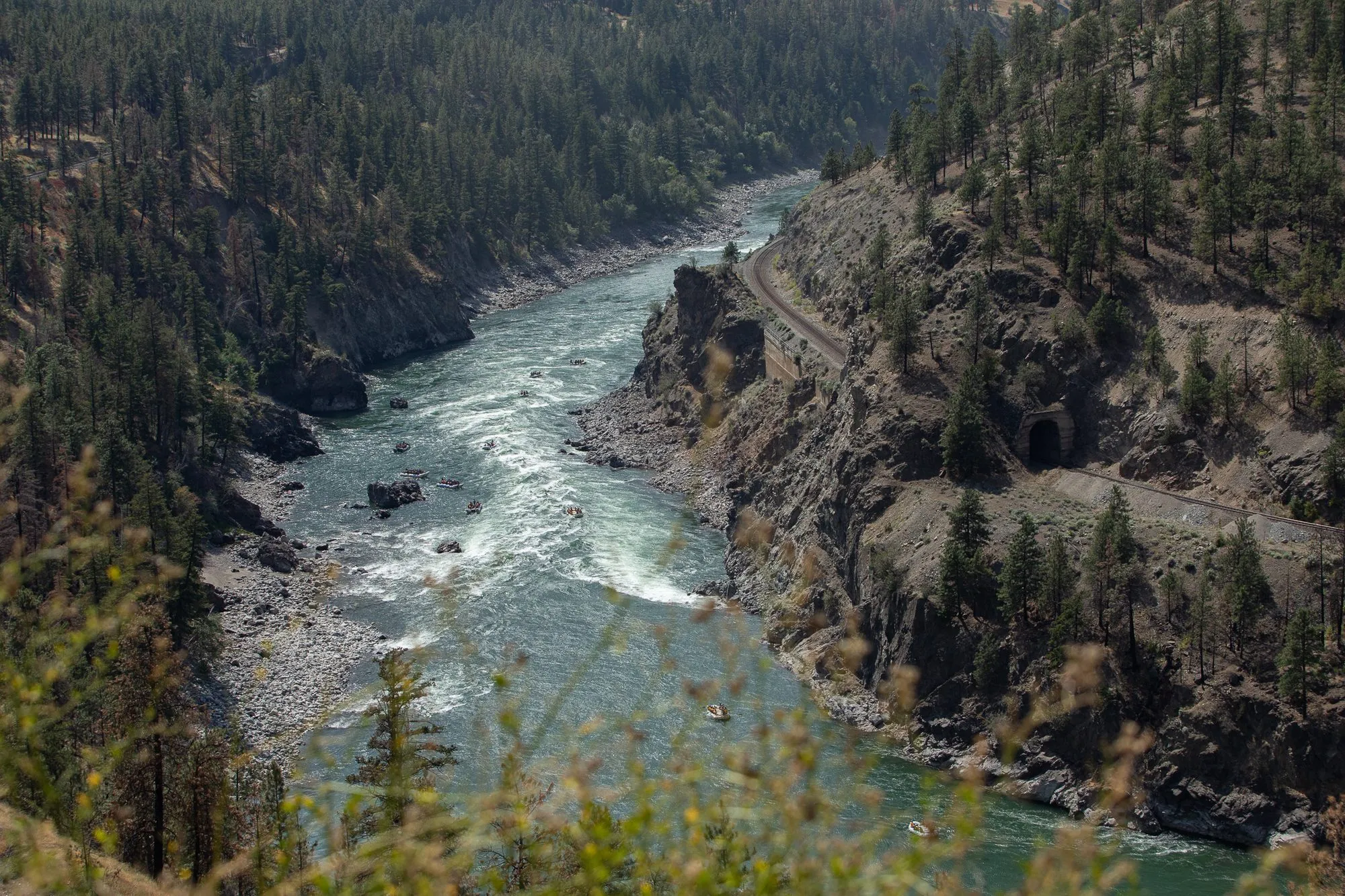 Looking down into the Thompson River canyon, with rapids in the river and a railway line and tunnel cut into the right bank.