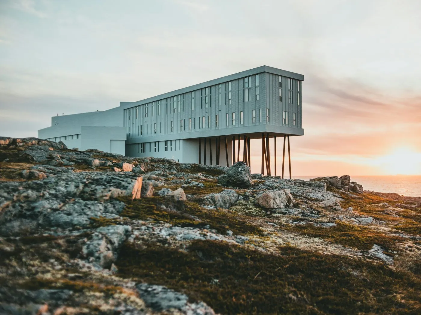 The Fogo Island Inn, a long white building with one end raised on thin steel legs, on bare rock at sunset.