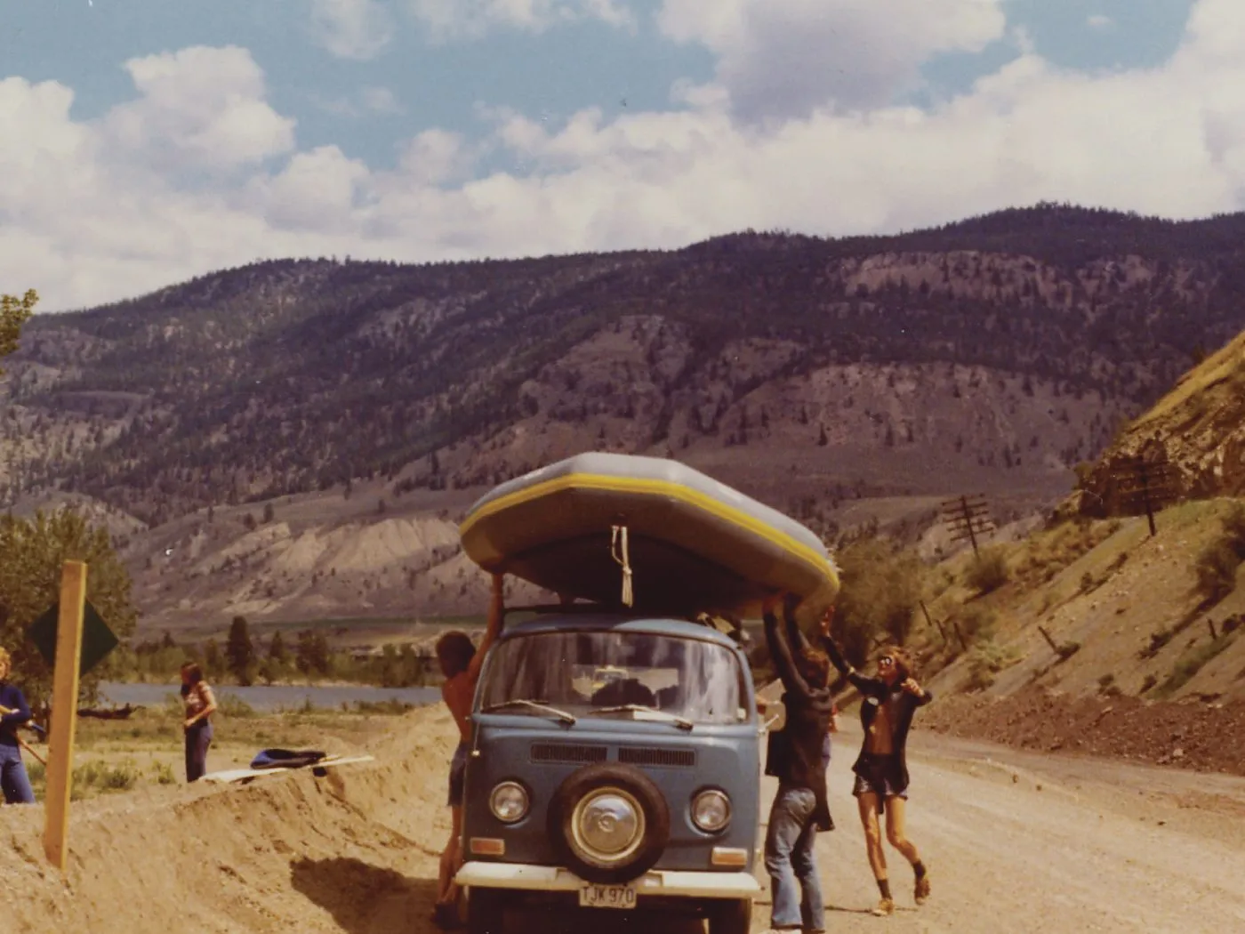 A 1970s Volkswagen van with a rubber raft on its roof, parked on a dirt road in dry hills while people unload it.