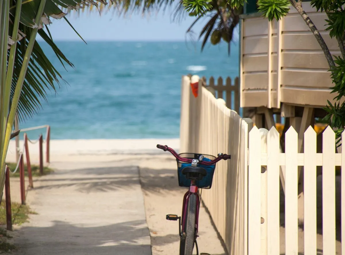 A bicycle with a basket leans on a cream picket fence beside a sandy path to the beach in Key West, with the sea beyond.