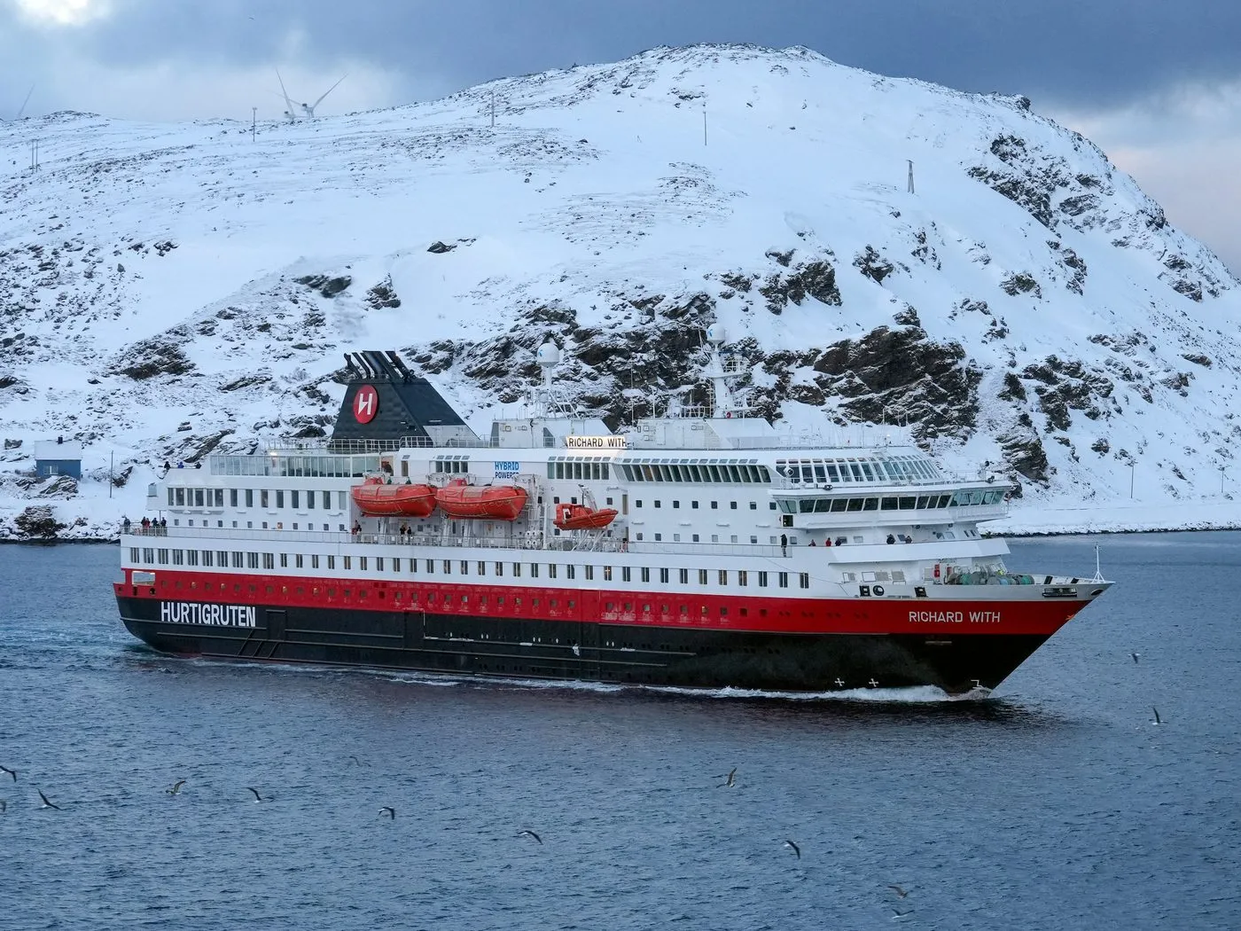 A red, white and black Hurtigruten ship passing a snow-covered mountain in northern Norway.