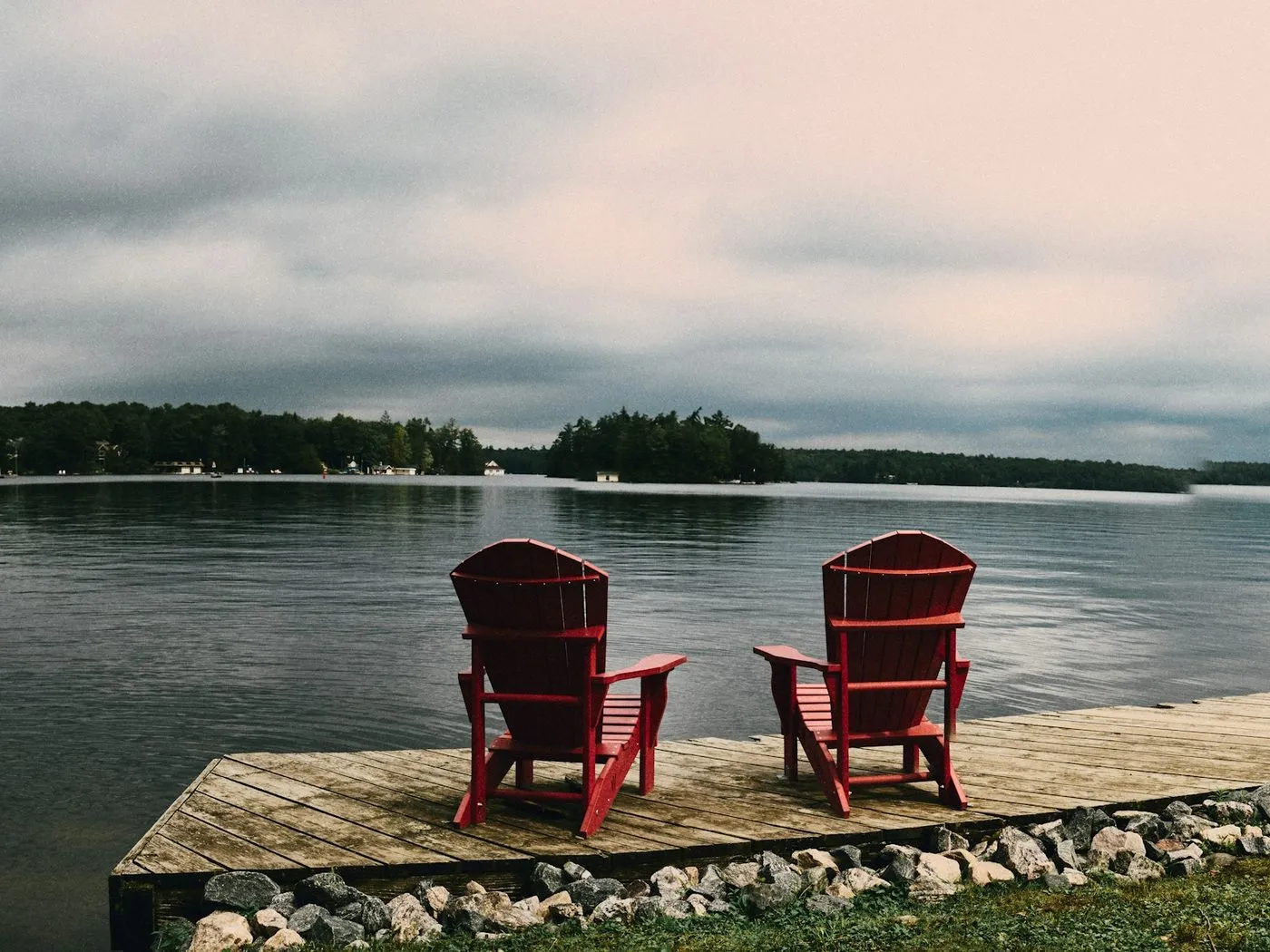 Two red Muskoka chairs side by side on a wooden dock, facing a calm lake with wooded islands.
