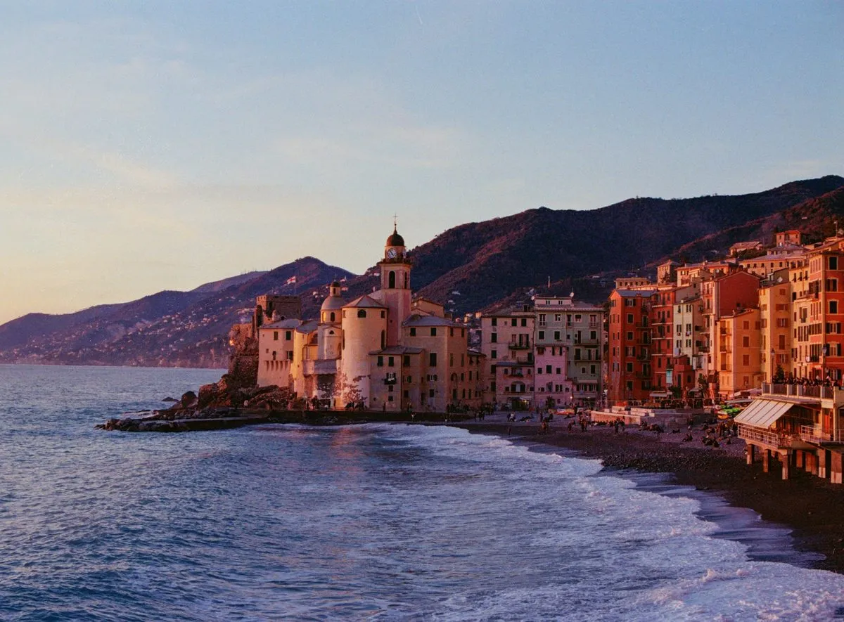 The tall painted houses and basilica of Camogli on the shore at sunset, with wooded hills behind.