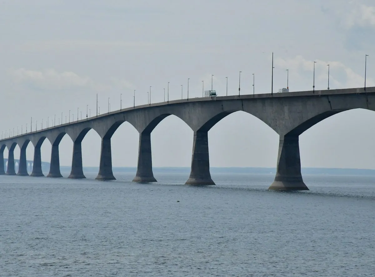 The Confederation Bridge curving away across the Northumberland Strait on tall concrete piers, under a hazy sky.