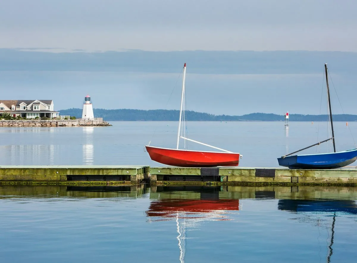 A red sailboat and a blue sailboat rest on a tidal dock at St. Andrews, with a white lighthouse across the still water.