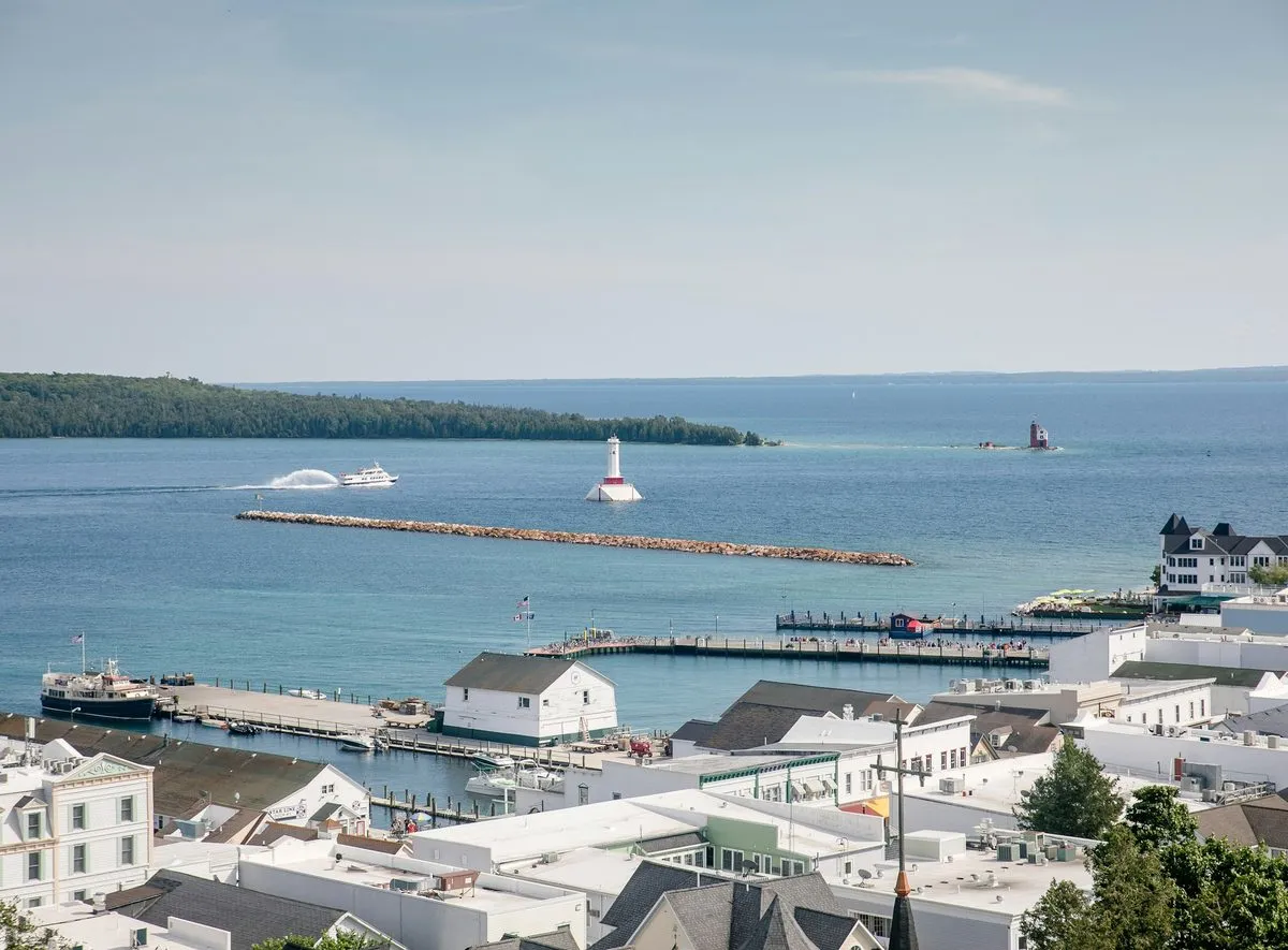 Looking down over the rooftops and marina of Mackinac Island to Lake Huron, with a white lighthouse offshore.