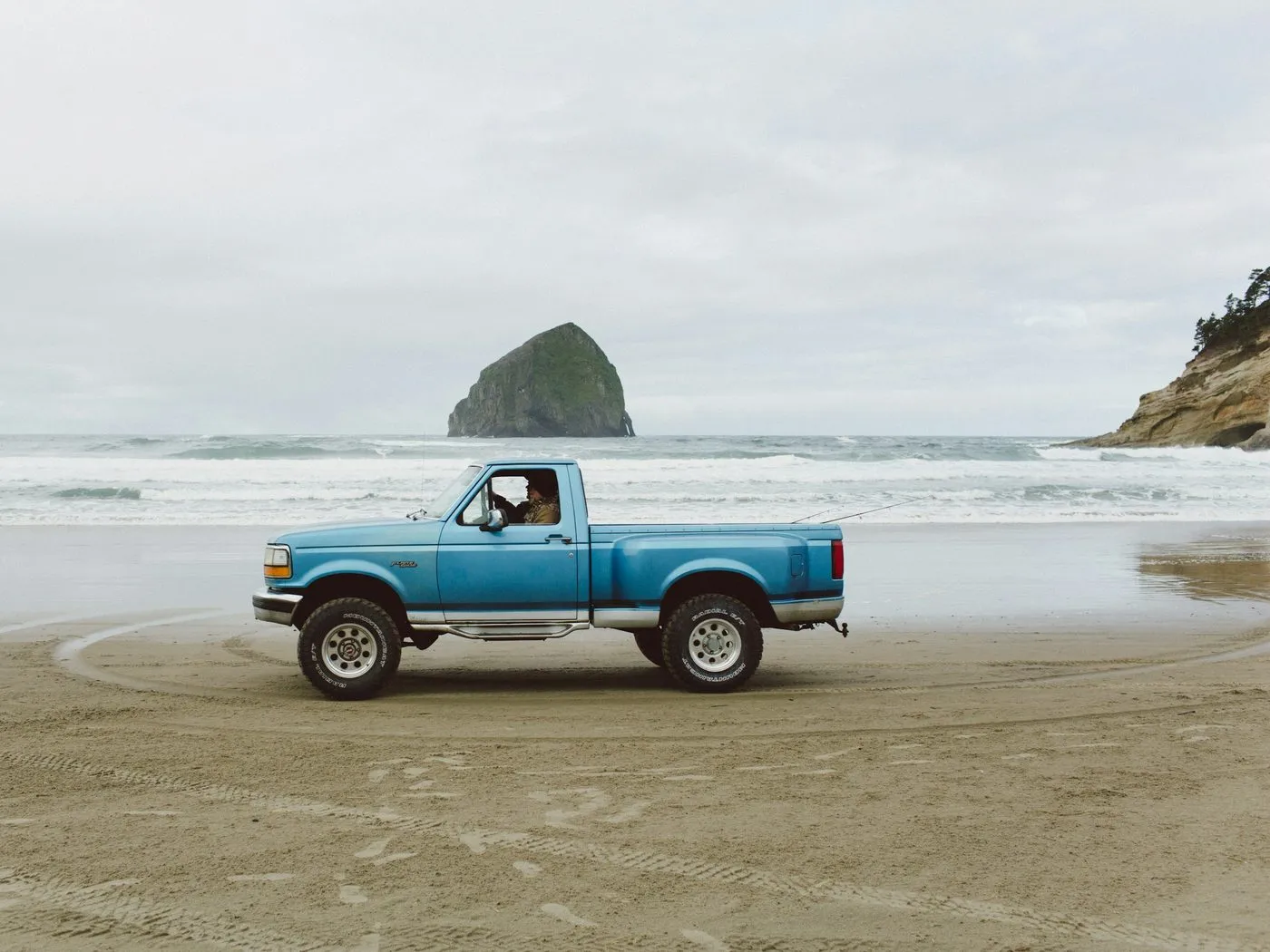 An old blue pickup truck on the wet sand at Pacific City, with Haystack Rock offshore under grey cloud.