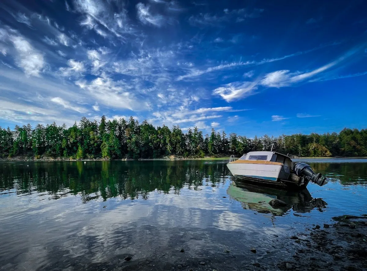 A small motorboat rests in still shallow water off Salt Spring Island, with a forested shore reflected under streaks of high cloud.