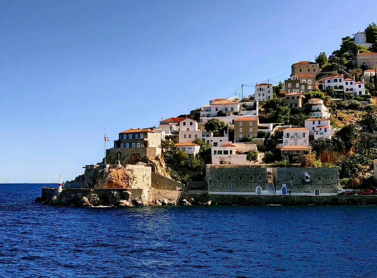Stone and whitewashed houses with terracotta roofs climb a rocky headland above deep blue sea on Hydra.