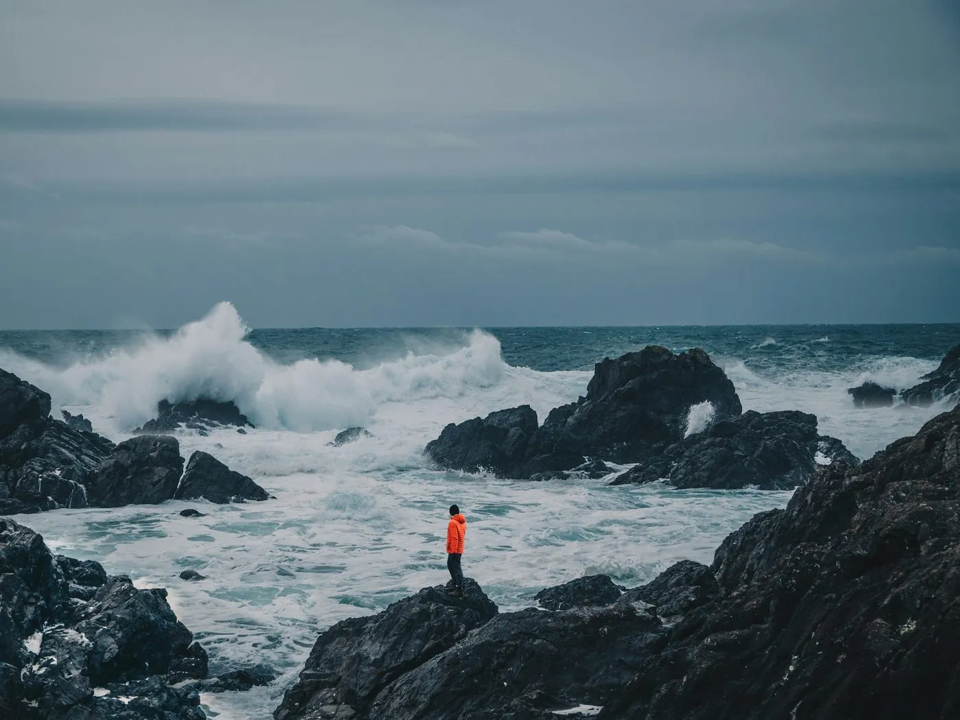A person in an orange jacket standing on dark rocks, watching large waves break in a grey sea near Tofino.