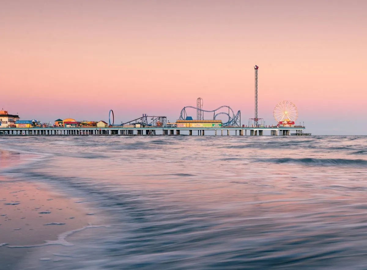 The Pleasure Pier at Galveston at dusk, its roller coaster and Ferris wheel lit against a pink sky above the surf.