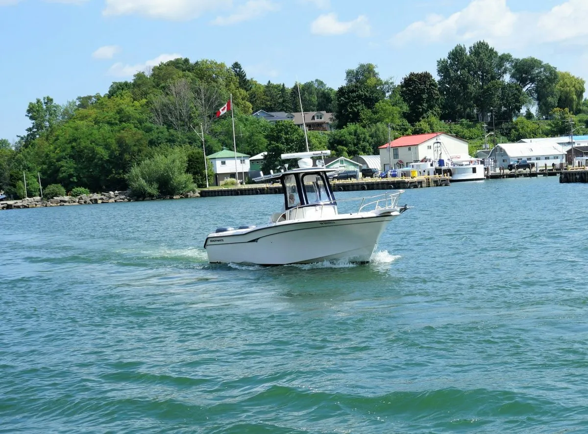 A white motorboat crosses the harbour mouth at Port Dover, with a Canadian flag, boathouses and trees along the shore behind it.