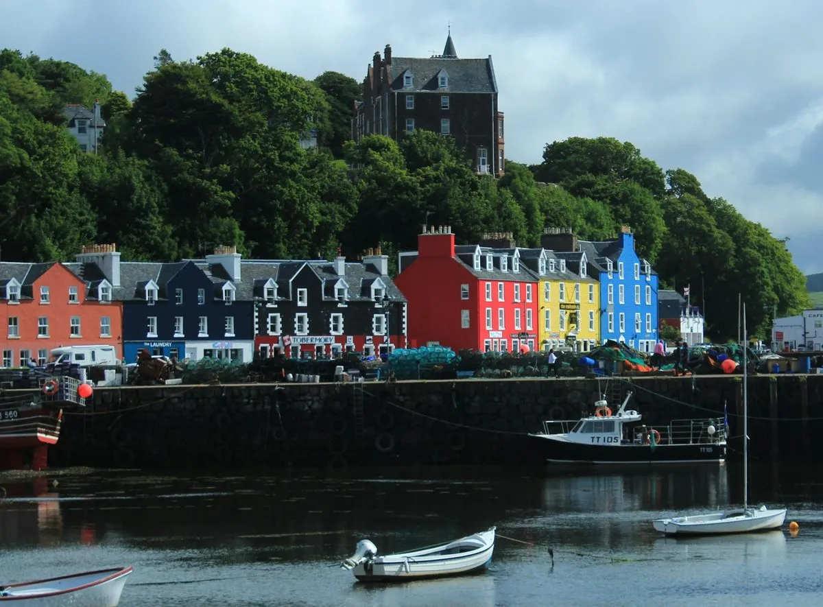 The painted houses of Tobermory’s Main Street in red, yellow, blue and black, above a stone harbour wall with fishing boats tied up below.