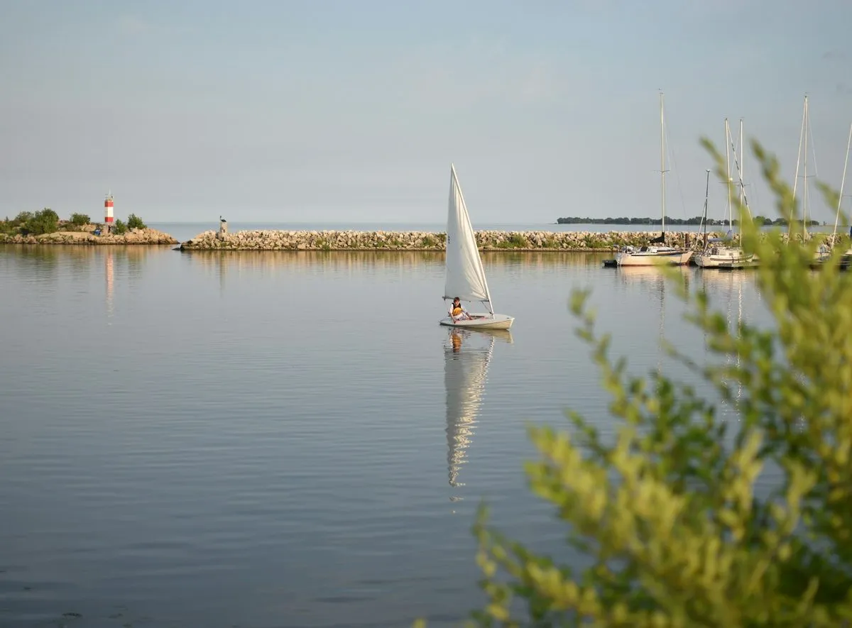 A small sailboat crosses the calm harbour at Gimli, with masts and a breakwater behind it.
