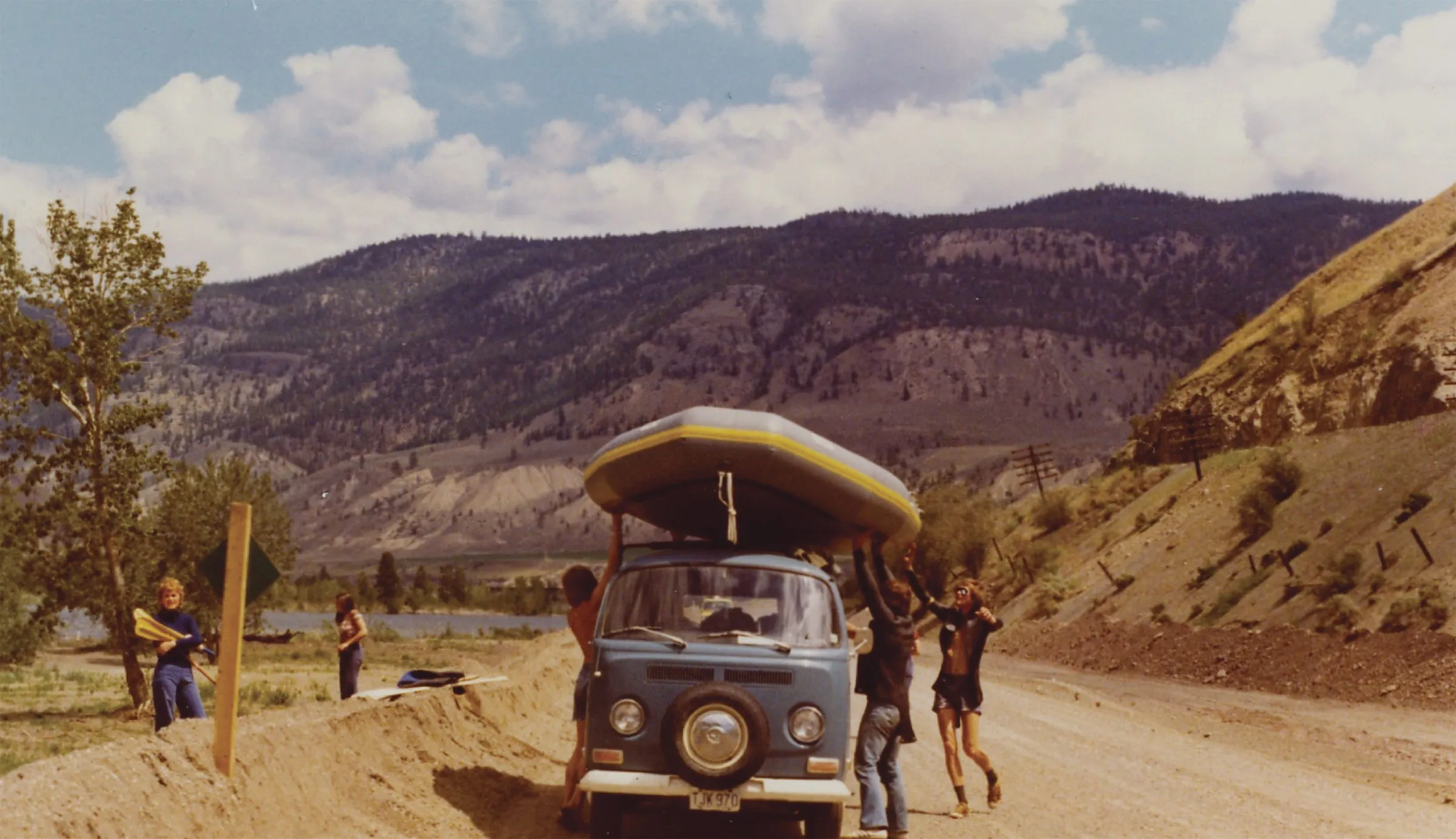 A 1970s Volkswagen van with a rubber raft on its roof, parked on a dirt road in dry hills while people unload it.