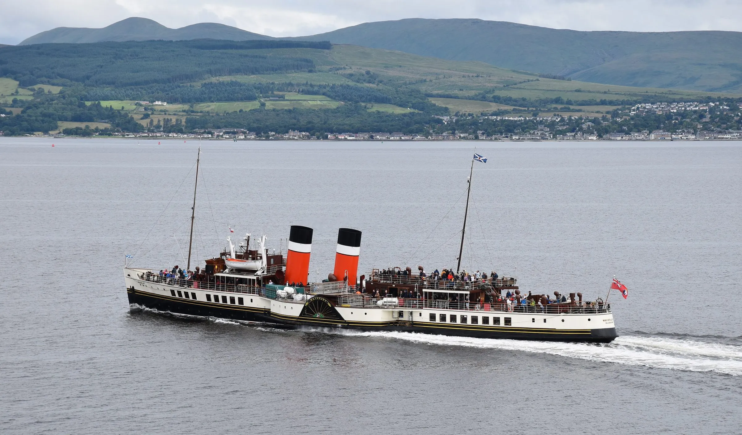 The paddle steamer Waverley crossing the Firth of Clyde, with two red, white and black funnels, crowded decks and green hills behind.
