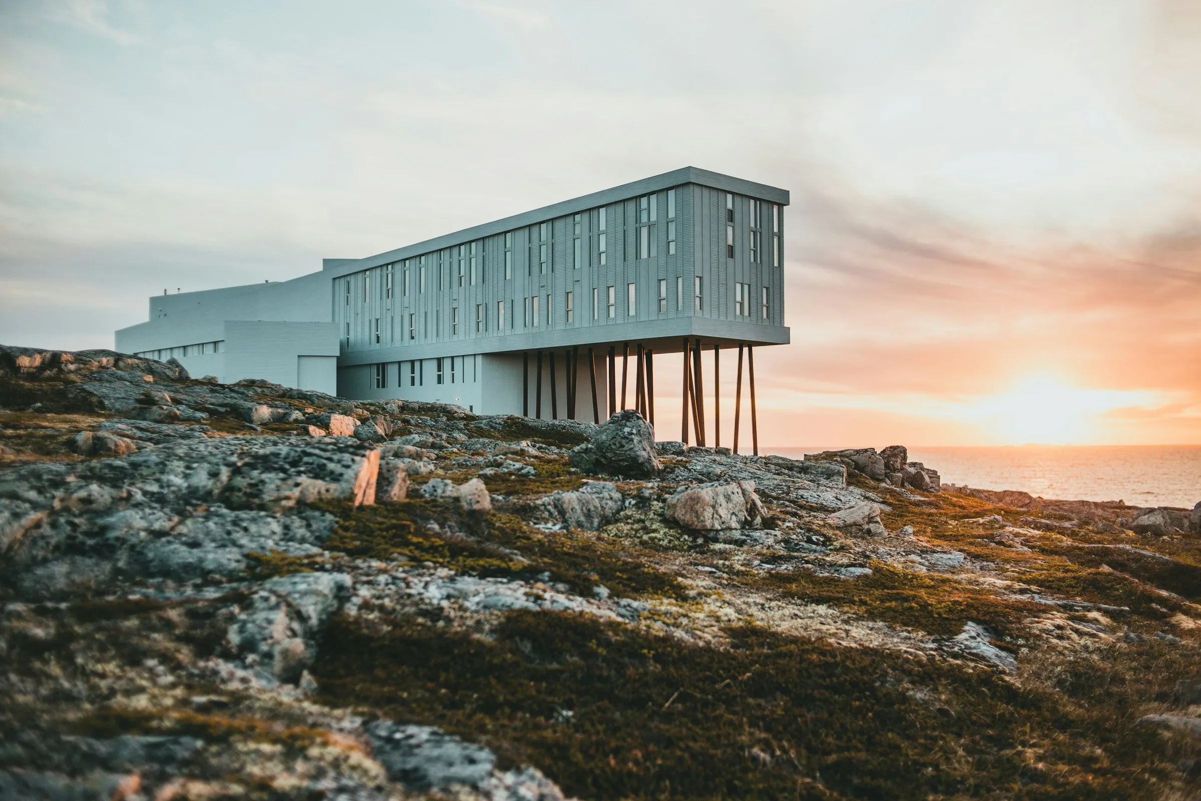The Fogo Island Inn, a long white building with one end raised on thin steel legs, on bare rock beside the sea at sunset.