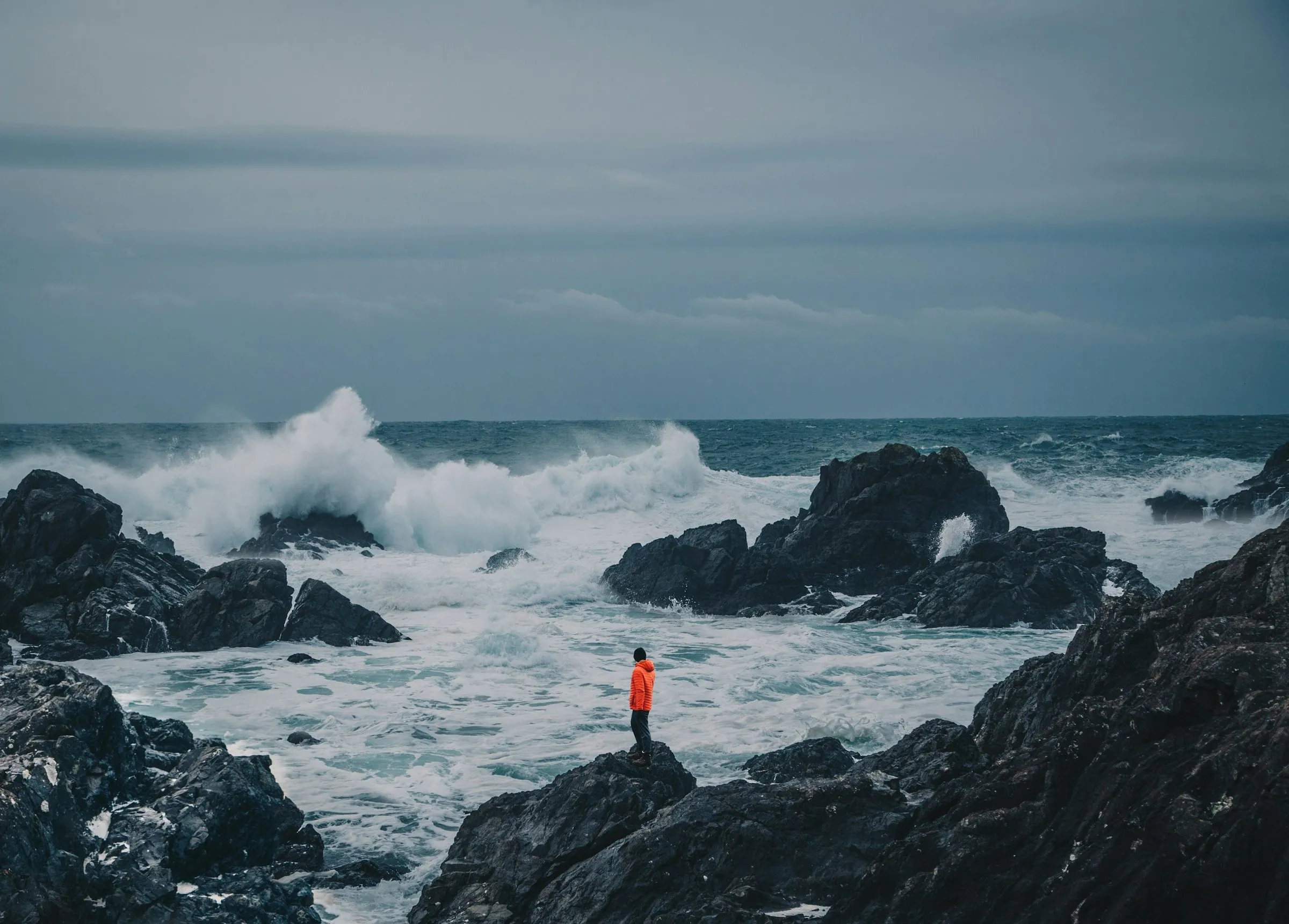 A person in an orange jacket standing on dark rocks, watching large waves break in a grey sea near Tofino.