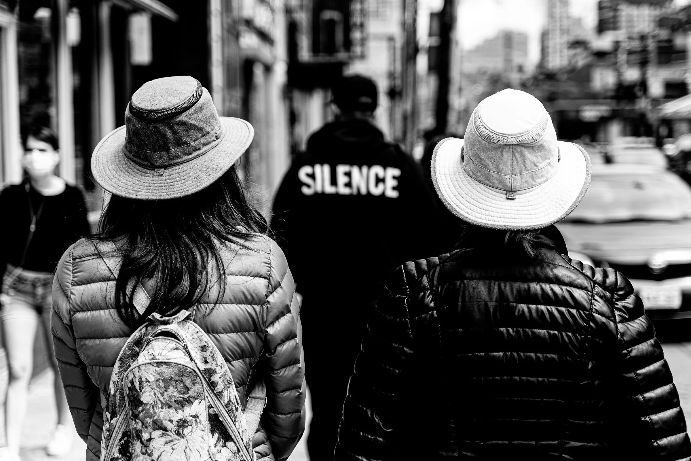 Black and white photo of two people seen from behind on a busy street, each wearing a wide-brimmed Tilley hat, with a third person between them whose jacket reads Silence.