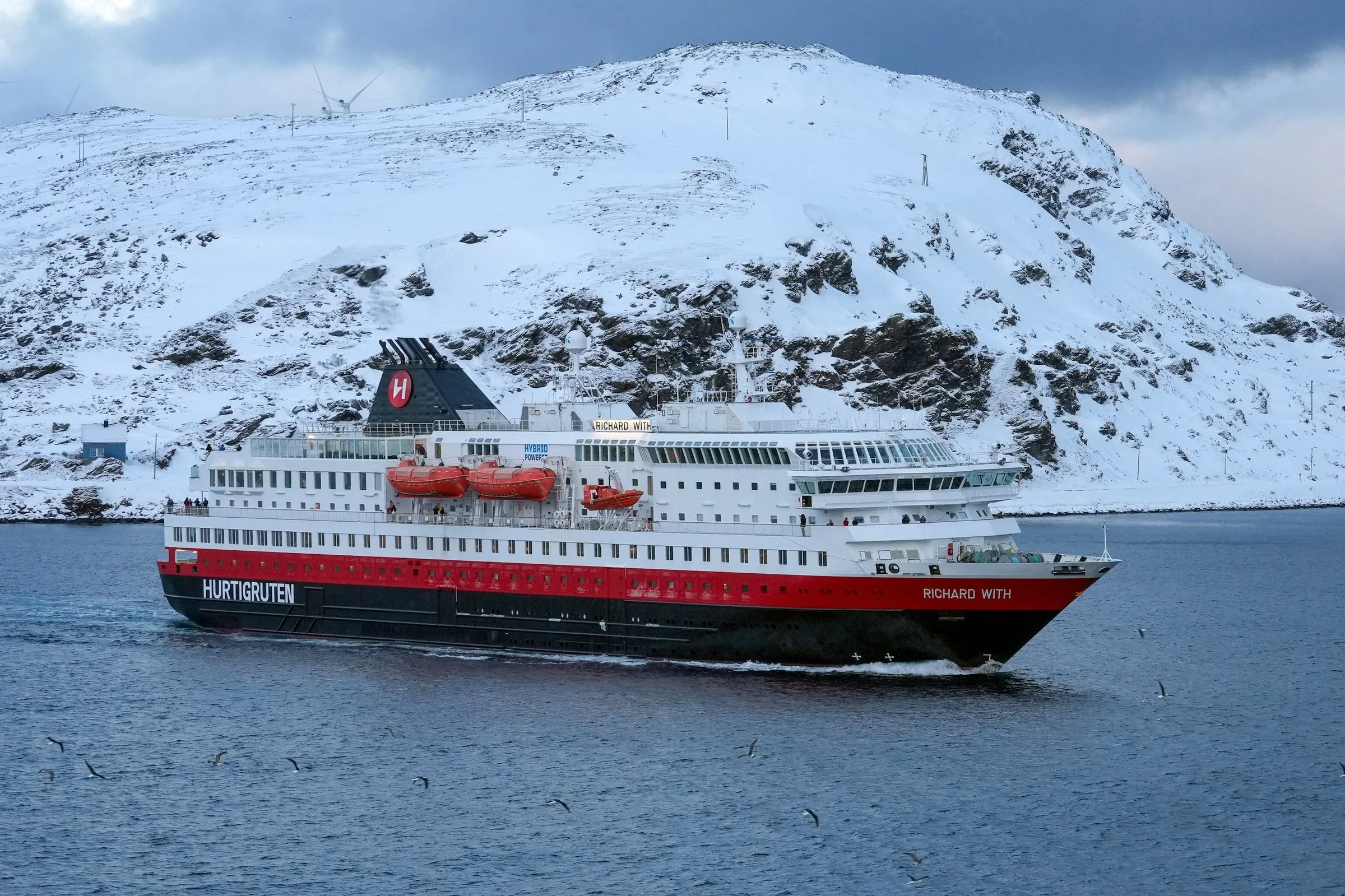 The Hurtigruten ship MS Richard With, red, white and black, passing a snow-covered mountain in Lofoten.