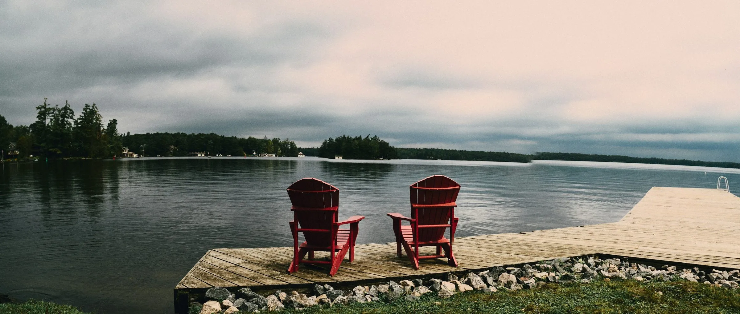 Two red Muskoka chairs side by side on a wooden dock, facing a calm lake with wooded islands under a pale evening sky.