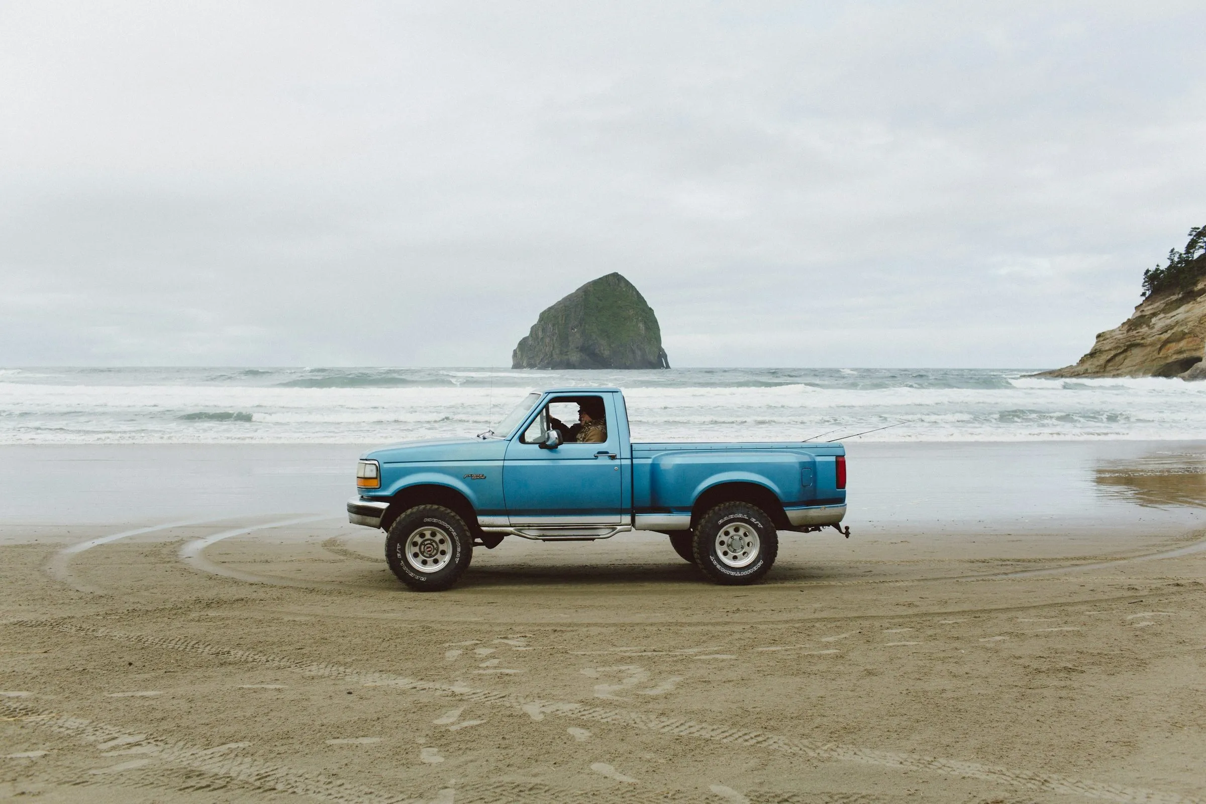 An old blue pickup truck on the wet sand at Pacific City, with Haystack Rock offshore under grey cloud.