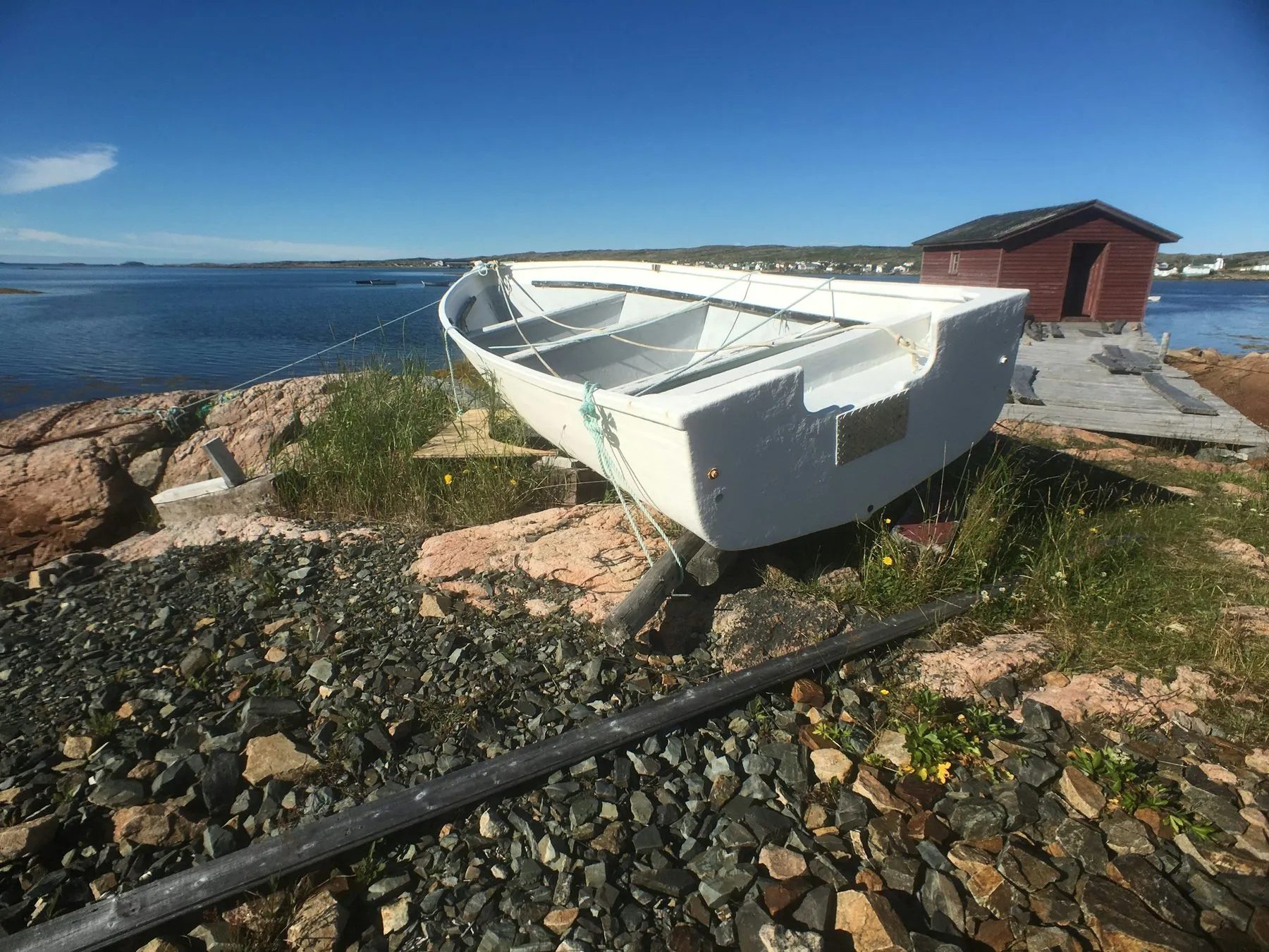 An old white wooden dory pulled up on rocks beside the sea, with a red fishing shed behind it.