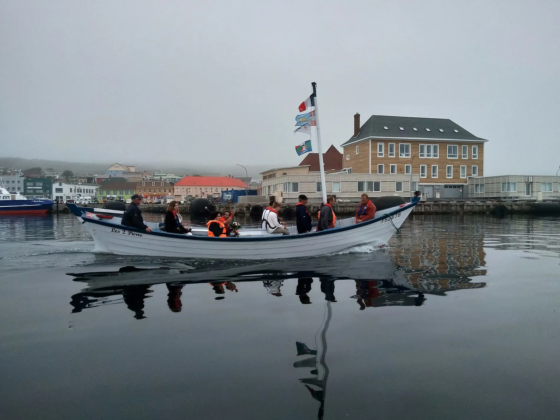 A white dory full of people crossing the still harbour in mist, with a stone building behind.