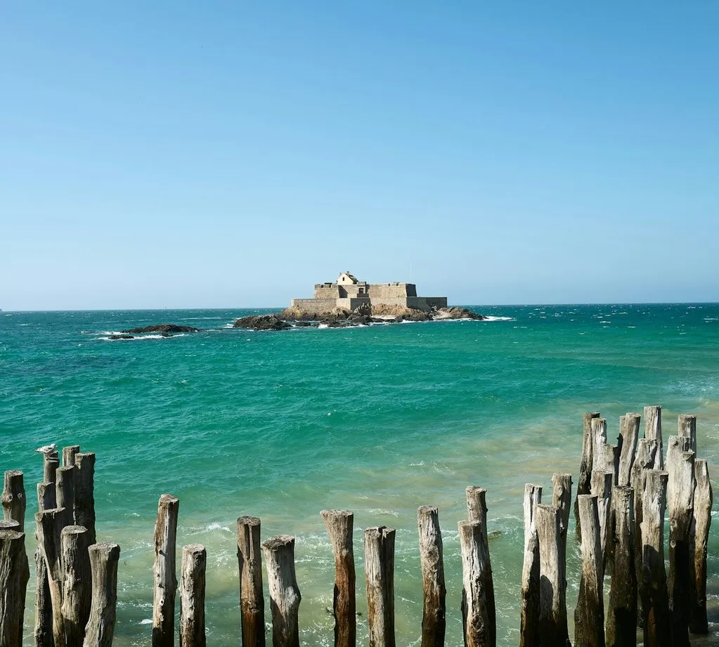 Fort National on its rock off Saint-Malo, across turquoise water, with rows of wooden breakwater posts in the foreground.