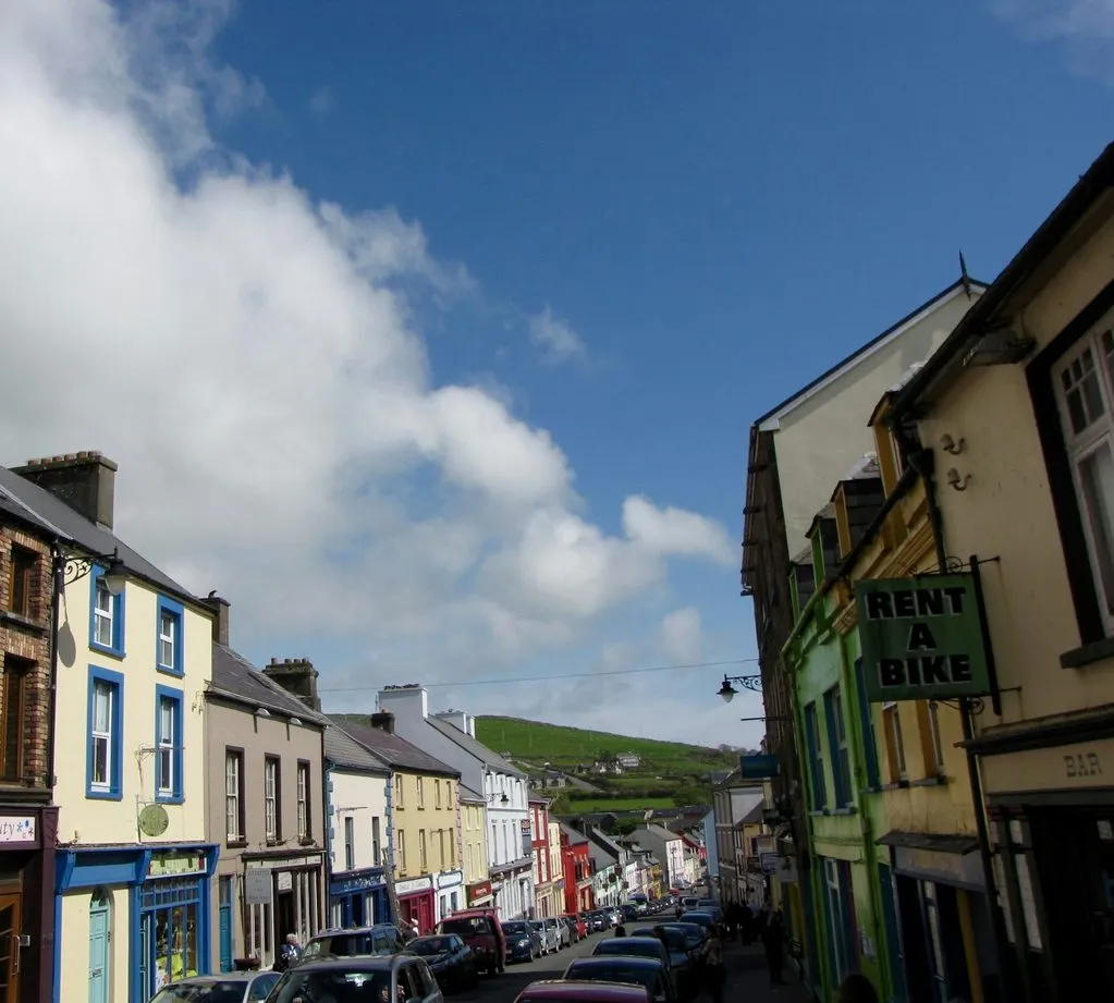 Looking down a street of painted shopfronts in Dingle, past a sign reading Rent a Bike, to green hills.