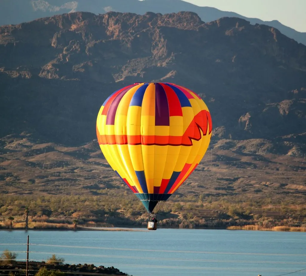 A striped hot-air balloon floats low over Lake Havasu, with bare desert mountains behind it.