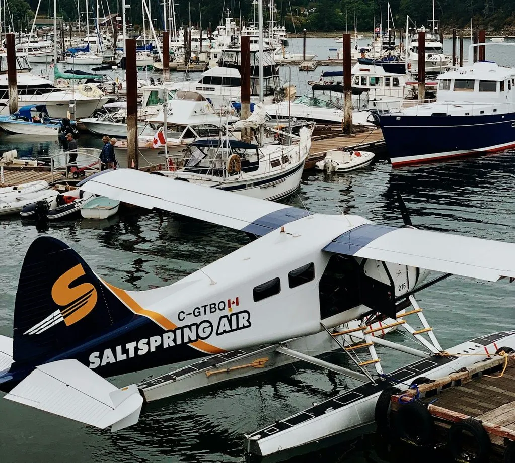 A Salt Spring Air float plane tied to the dock in Ganges Harbour, with sailboats and cruisers moored in the marina behind it.