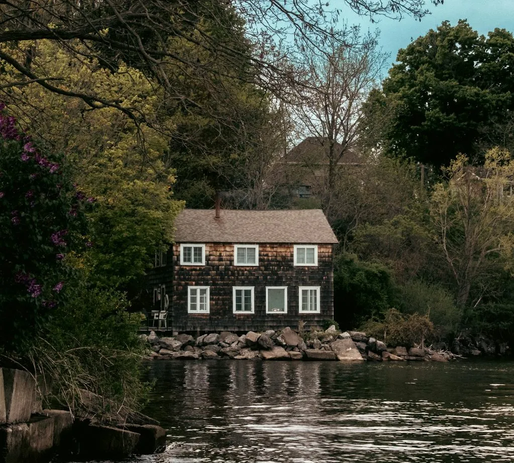 A weathered shingled boathouse with white-trimmed windows sits on the rocks at the river’s edge in Port Dover, under tall trees.