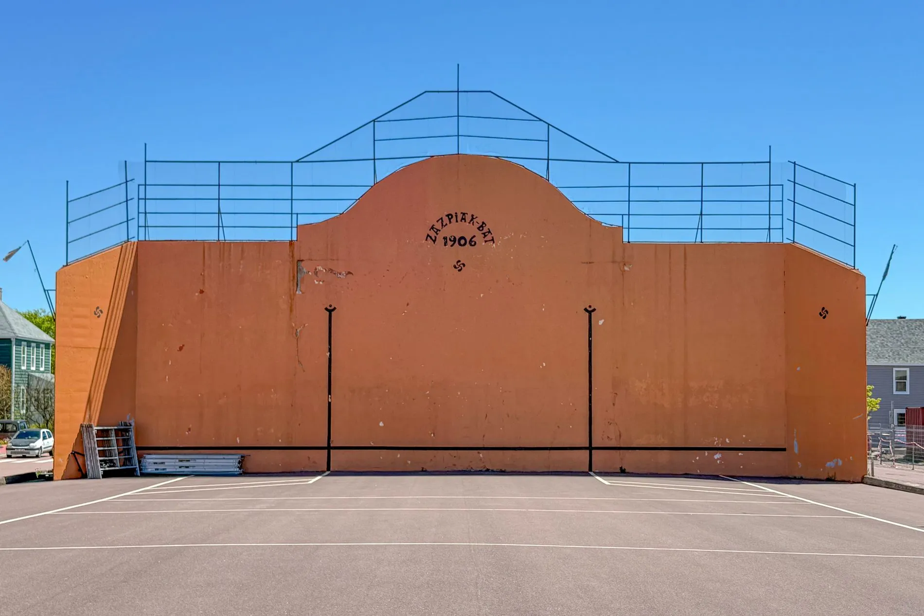 A tall orange wall with a curved top, standing at the end of a paved court under a blue sky.