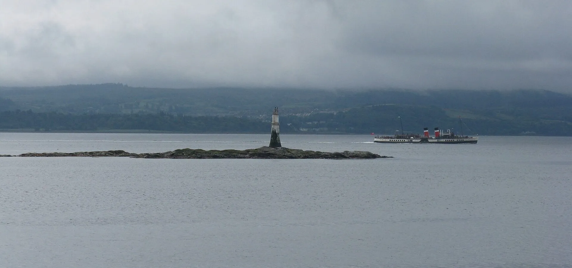 A low reef with a small beacon on it in grey water, with Waverley passing in the distance under heavy cloud.