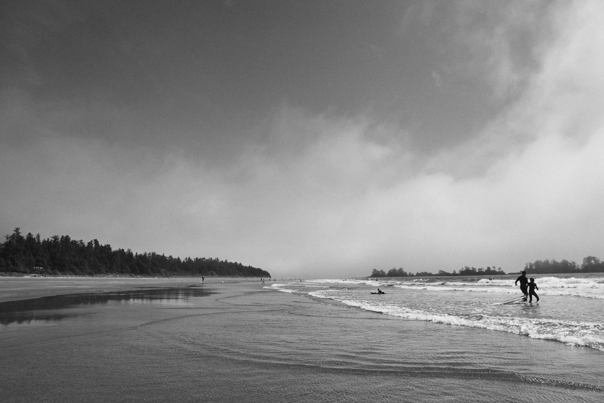 A black and white view of a wide beach near Tofino under heavy cloud, with two surfers carrying boards into the waves.