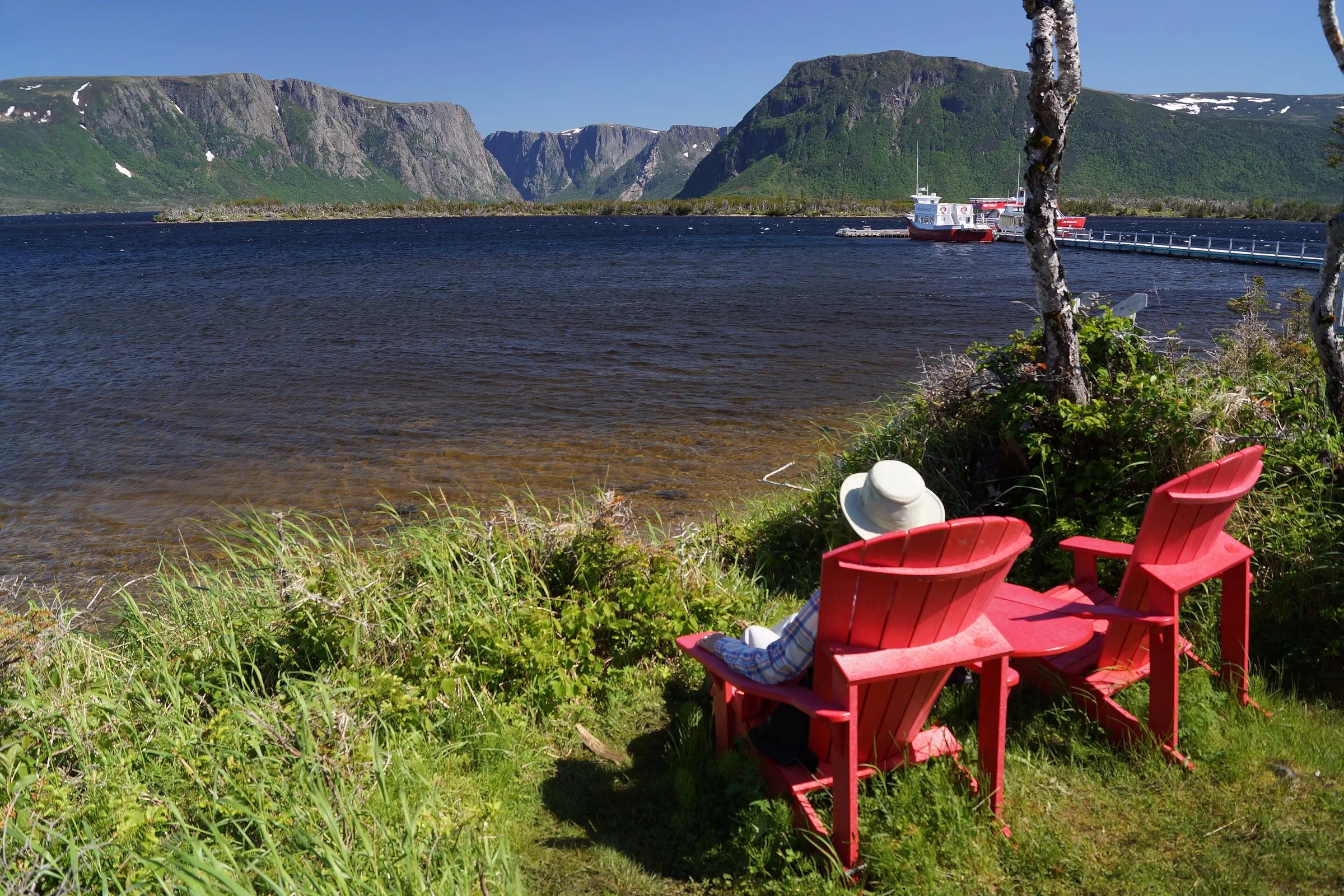 Two red chairs on a grassy bank above Western Brook Pond in Gros Morne National Park, one occupied by a person in a white sun hat, with flat-topped cliffs across the water.