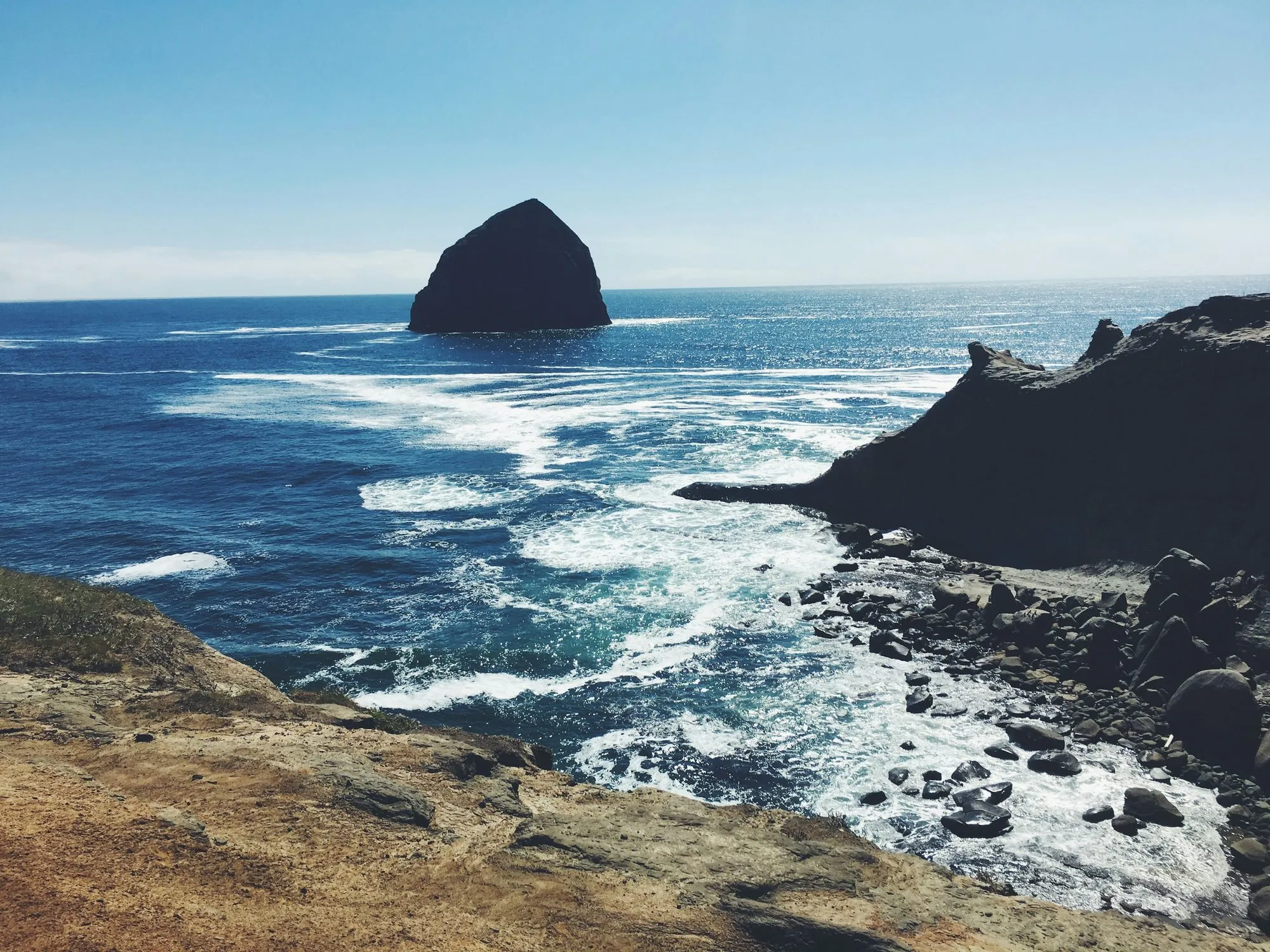 Haystack Rock, a dark basalt sea stack, seen across white surf from the rocks of Cape Kiwanda.