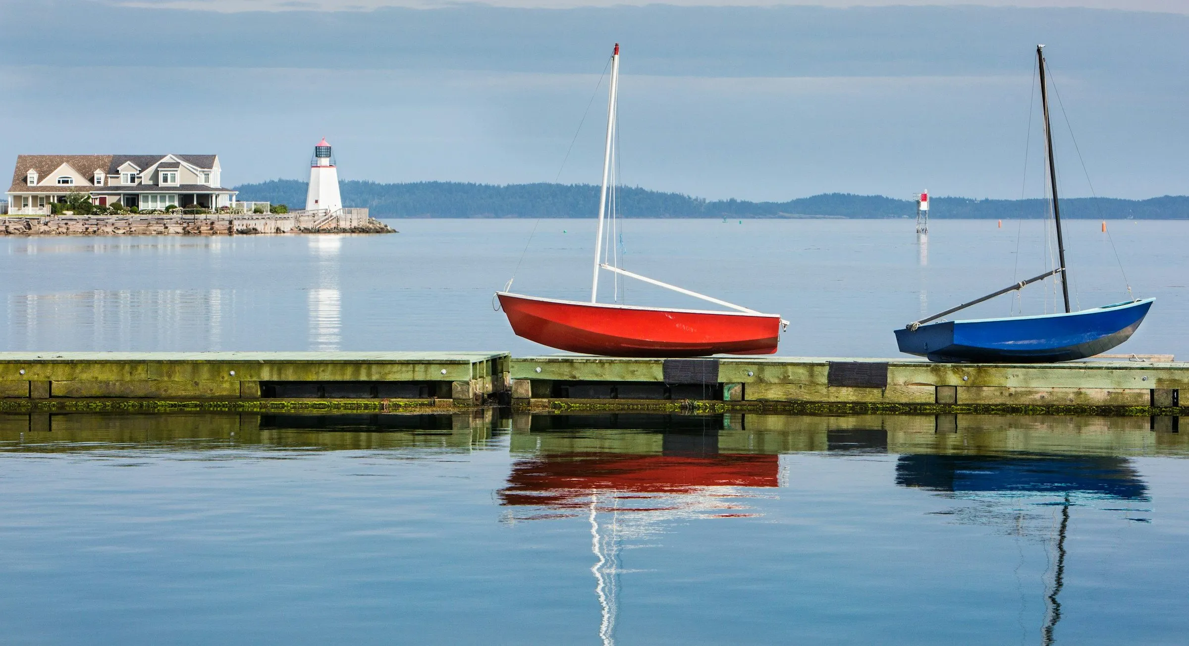 A red sailboat and a blue sailboat rest on a tidal dock at St. Andrews, with a white lighthouse across the still water.