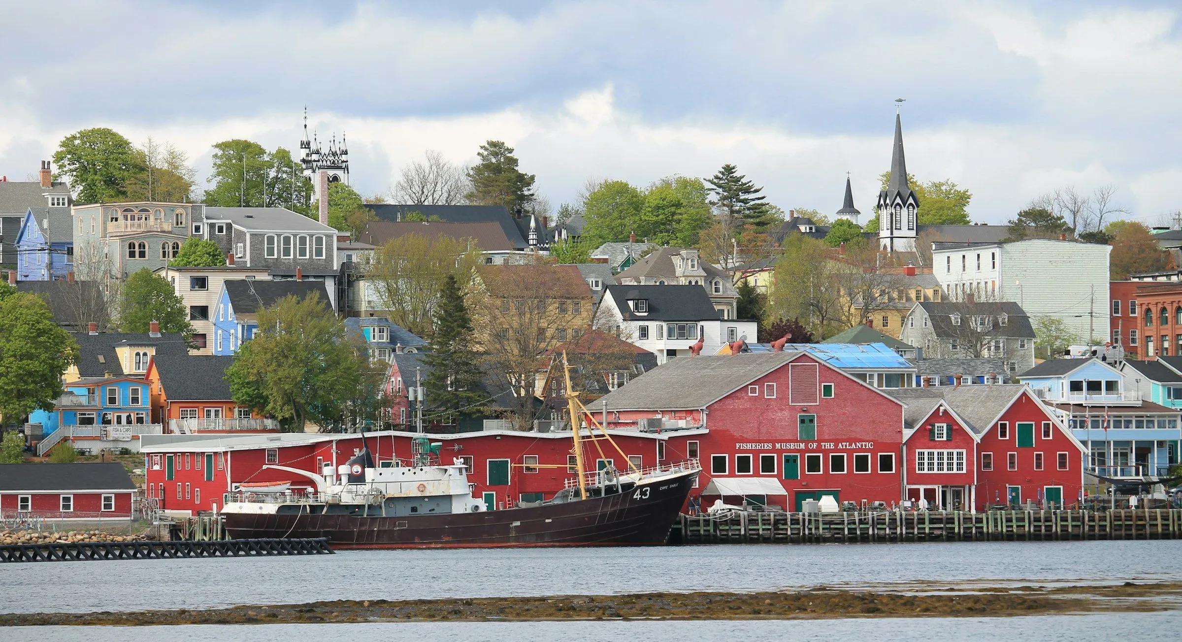 The red wharf buildings of the Fisheries Museum of the Atlantic on the Lunenburg waterfront, with a fishing vessel alongside and the town rising behind.