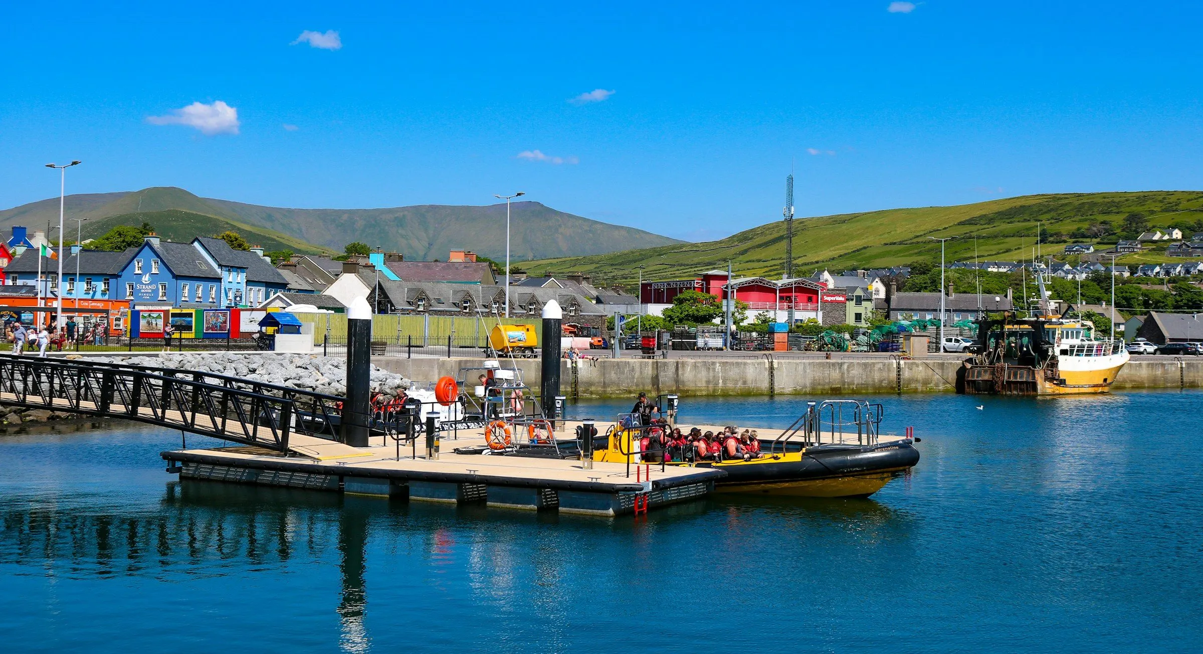 Tour boats tied to a pontoon in Dingle harbour, with painted buildings and green hills behind.