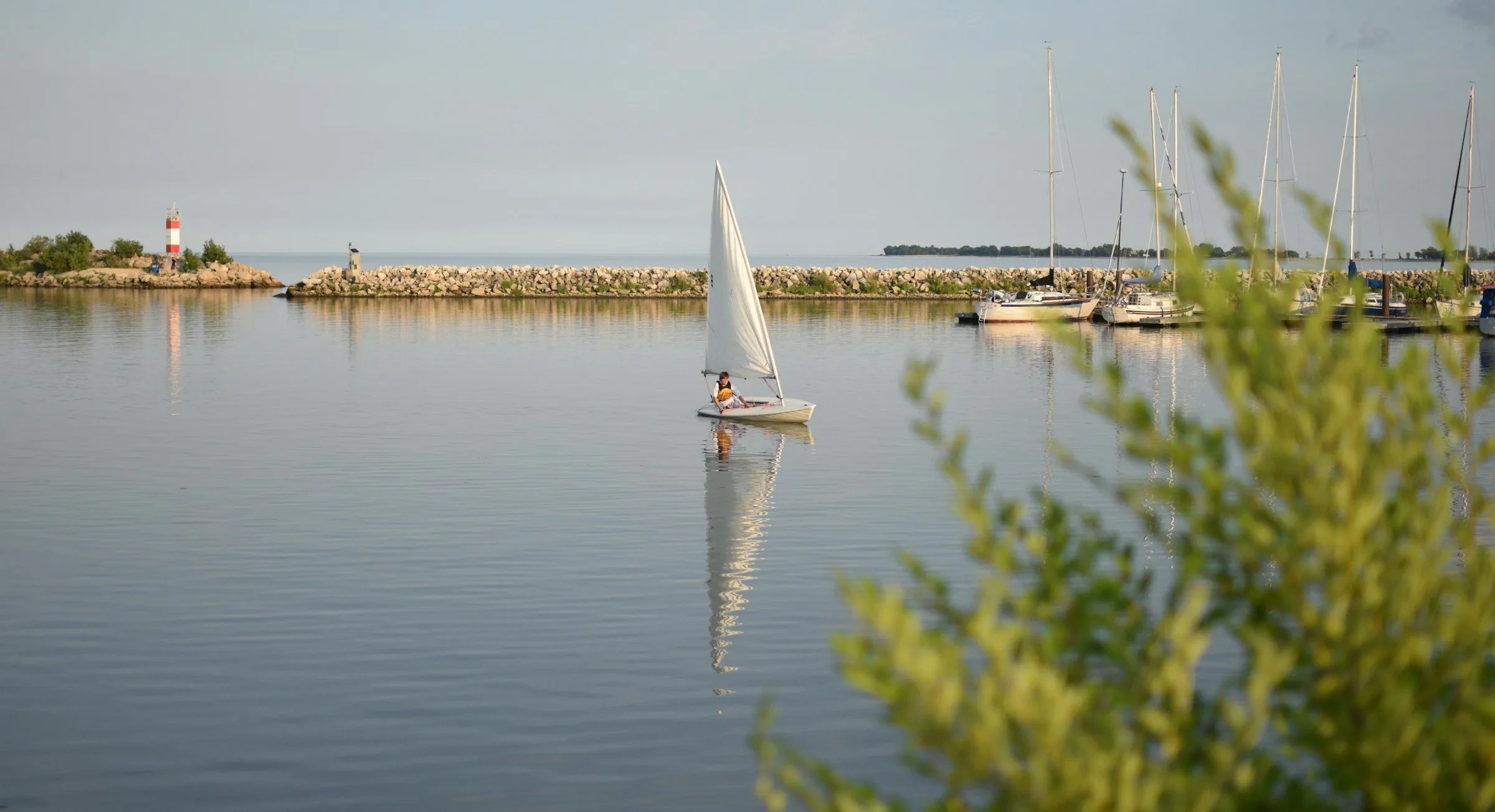A small sailboat crosses the calm harbour at Gimli, with masts and a breakwater behind it.