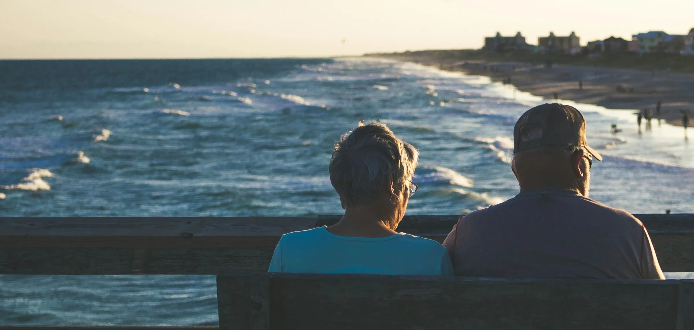 An older couple sit close together on a seaside bench, watching the waves roll in at golden hour.