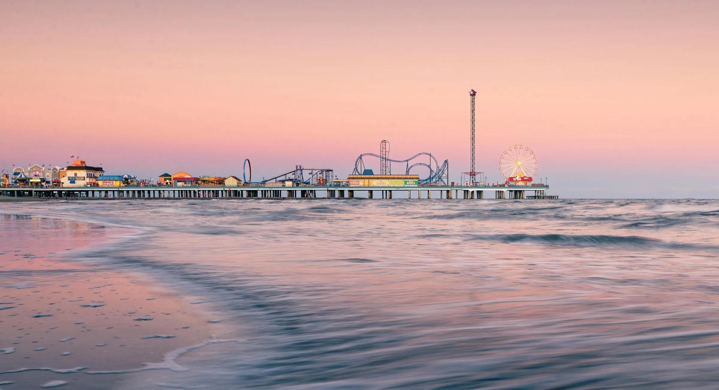 The Pleasure Pier at Galveston at dusk, its roller coaster and Ferris wheel lit against a pink sky above the surf.