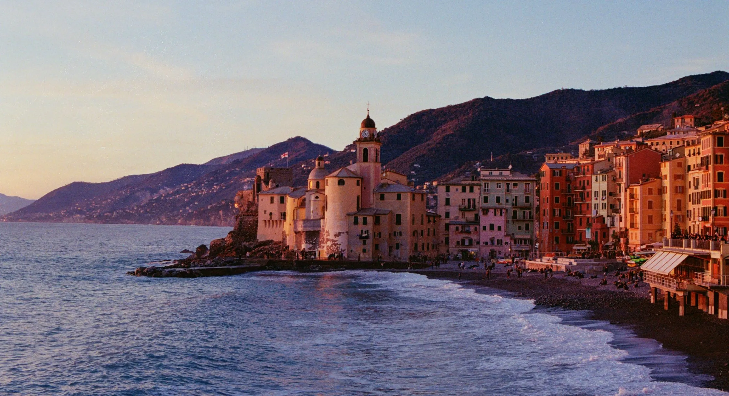 The tall painted houses and basilica of Camogli on the shore at sunset, with wooded hills behind.