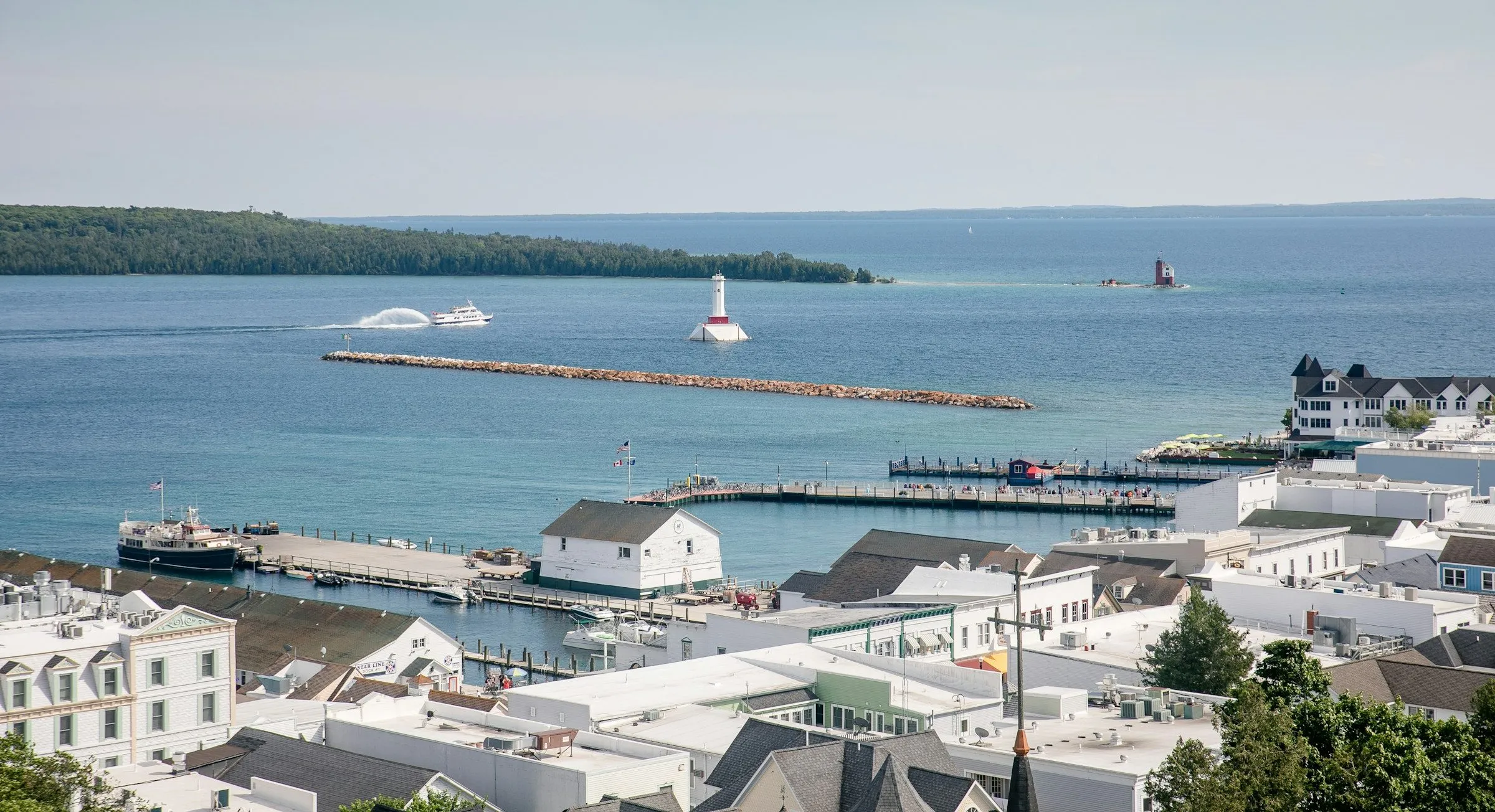 Looking down over the rooftops and marina of Mackinac Island to Lake Huron, with a white lighthouse offshore.