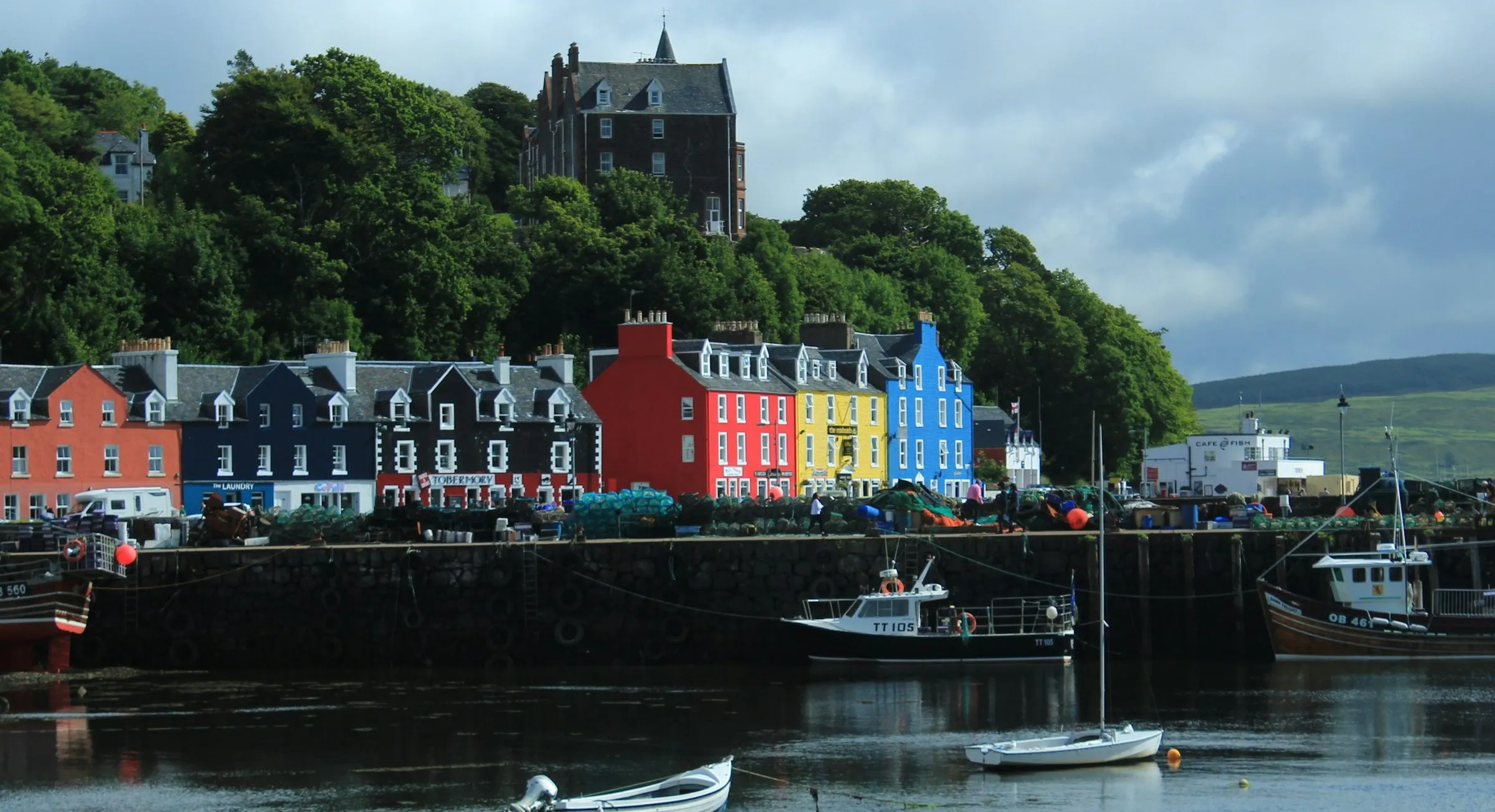 The painted houses of Tobermory’s Main Street in red, yellow, blue and black, above a stone harbour wall with fishing boats tied up below.